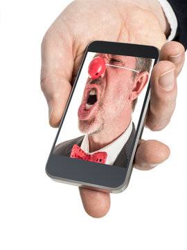 Closeup photograph of a man's hand holding a smart phone. On the screen of the smart phone is an angry clown in a business suit with a red rubber nose.