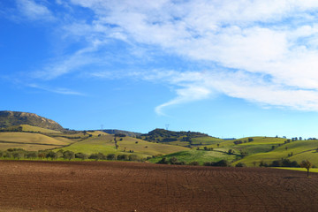 Fototapeta na wymiar Paesaggio con terreno arato in primo piano, delle colline sullo sfondo e le nuvole nel cielo azzurro