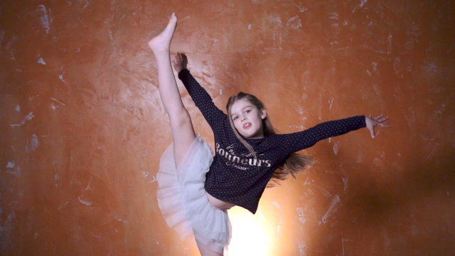 Flexible little girl doing vertical split in studio. Girl gymnast doing the splits isolated over orange modern background.