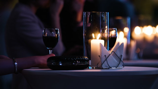 Close-up footage of elegantly decorated table at restaurant, woman holding a glass of red wine, fancy black clutch bag is near. Man brings a plate with food and have a drink.  