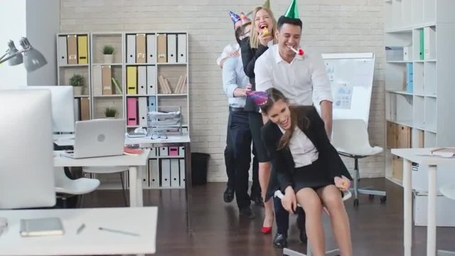 Group of office workers in birthday hats pushing chair with their colleague sitting on it, dancing and laughing