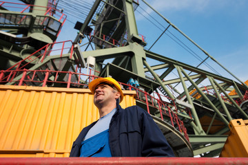 Smiling and happy electrical engineer standing and is looking at the side. In the background is a huge drill machine for coal digging. View from below. Photographed with wide angle lens.