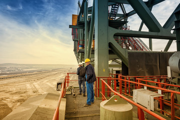 Two engineers standing on a huge drill machine and looking down at a mining site in distance. 