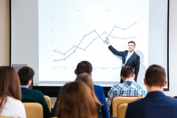 Smiling speaker standing and explaining graphs on business conference