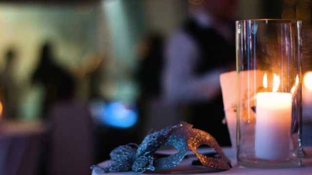 Masquerade gold venetian mask lies on a table in a nightclub during a costume party, white candle burning in a glass cup,  blurred footage of people partying on a background.