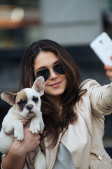 Beautiful young girl smiling and taking a selfie with her cute French bulldog puppy. Urban scene. Selective focus on girl.