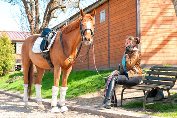 Woman riding trainer near chestnut horse speaking on mobile phon