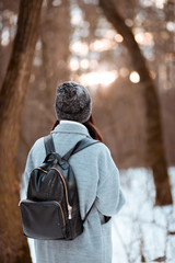 Portrait of a happy beautiful girl with brown hair in  winter forest.Attractive brunette woman dressed in a hipster style with black stylish leather backpack wearing grey coat and white pullover.