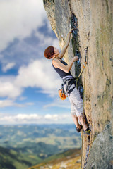 Young female rock climber climbing with rope and carbines an overhanging cliff against blue sky and mountains background. Summer time.