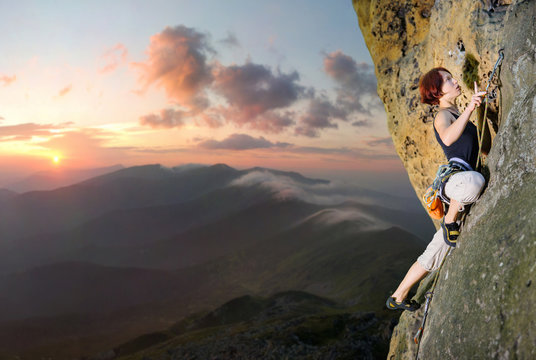 Athletic female rock climber climbing challenging route with rope and carbines on rocky wall against scenic sunset background. Summer time.
