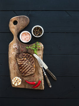 Grilled ribeye beef steak with herbs and spices on walnut cutting board over black wooden background