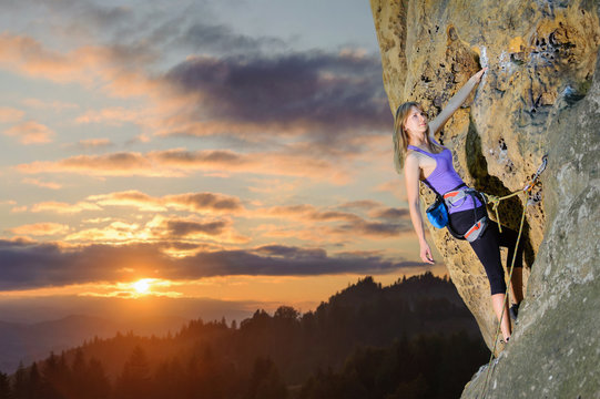 Young athletic girl climbing on big boulder with rope and carbines, against scenic sunset background. Hanging on one hand. Summer time.