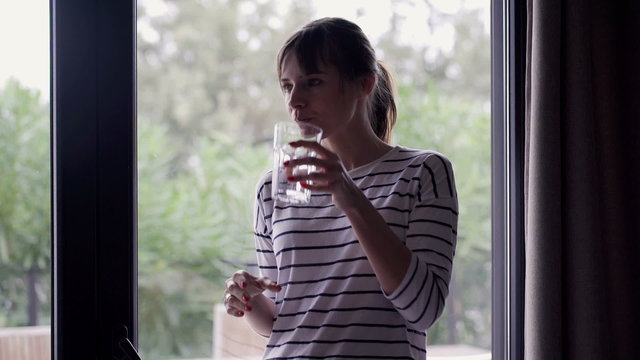 Young, pensive woman drinking water and looking by the window
