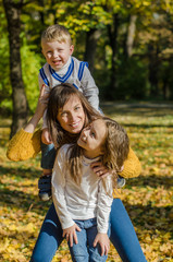 Fototapeta na wymiar Smiling mother hugs daughter and holds son on her shoulders in the autumn park