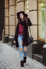 Fototapeta na wymiar Outdoor full body portrait of a young beautiful woman walking at the street. Model wearing stylish black hat and clothes. Elegant outfit. Female fashion. City lifestyle. Toned