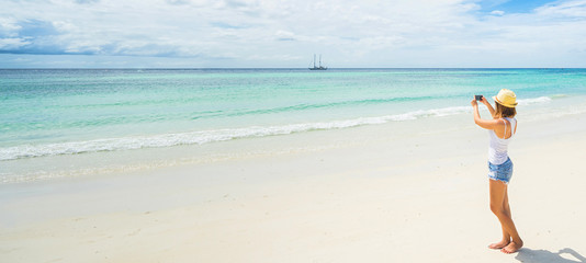 Fototapeta na wymiar Female tourist taking photo of beautiful island beach on smartphone. Banner edition.