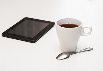 White cup of tea, digital tablet and spoon on light wooden desk.