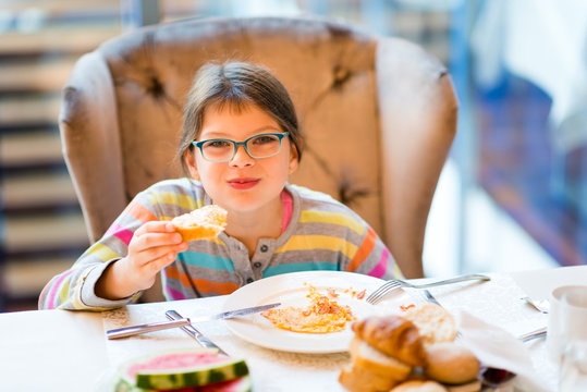 Happy girl in glasses eating breakfast - egg omelet, toast, watermelon and tea - sitting in comfortable armachair