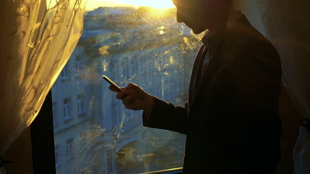 Handsome man using phone, smiling against the background of a window, curtains 