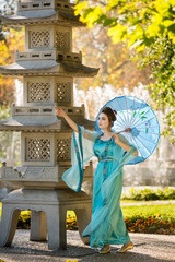 Fototapeta premium beautiful geisha with a blue umbrella near stone pagoda on background of blur fountain