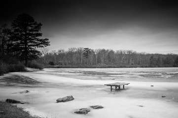 Black and white winter scene of picnic table in frozen lake in desolate forest 