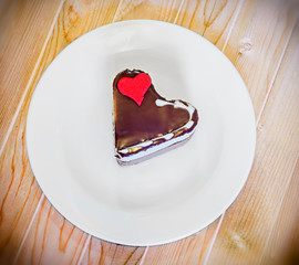 Heart shape chocolate cake with sugare red heart on white plate, wood background, close up, isolated.