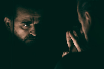 Angry man looking at his reflection in a mirror, in a dark and low light room