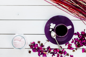 cup of tea or coffee on violet plate, silver spoon, marshmallow in glass vase, lump sugar, violet purple dry decor scattered on white colored wooden table,  top view