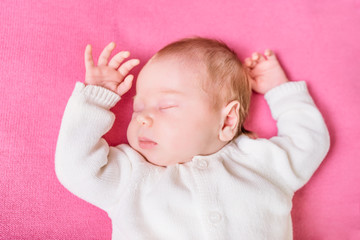 2 week old baby with closed eyes wearing knitted white clothes lying on pink plaid. Sweet little baby sleeping on pink sofa. Security and childcare concept. Selective focus on eye