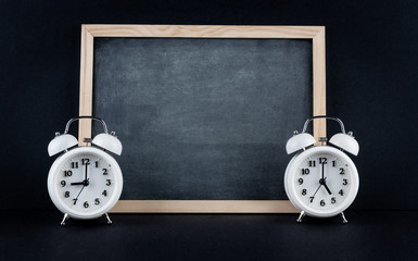 Two vintage alarm clocks showing 9 and 5 o'clock with empty chalkboard on black background. Nine to...