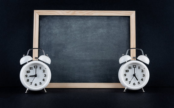 Two vintage alarm clocks showing 9 and 5 o'clock with empty chalkboard on black background. Nine to five corporate working hours concept.
