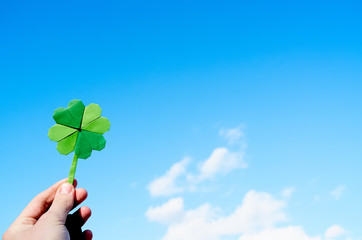 Hand holding green paper origami folded shamrock on blue sky background. Sunny weather outdoors....