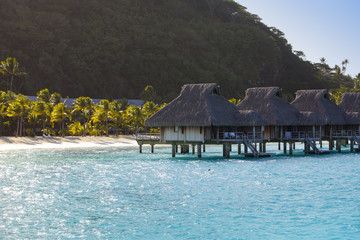 Typical Polynesian landscape - island with palm trees and small houses on water in the ocean and mountains on a background