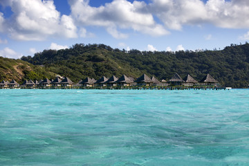 Typical Polynesian landscape - island with palm trees and small houses on water in the ocean and mountains on a background