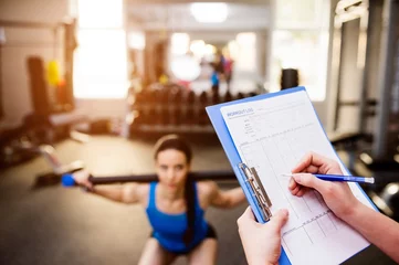 Tuinposter Woman exercising in gym, personal trainer, plan on clipboard © Halfpoint
