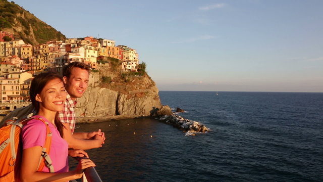 People looking at sunset on holidays travel talking. Young backpacking Asian woman and Caucasian man enjoying ocean view. Young people on vacation in Manarola, Cinque Terre, Liguria, Italy
