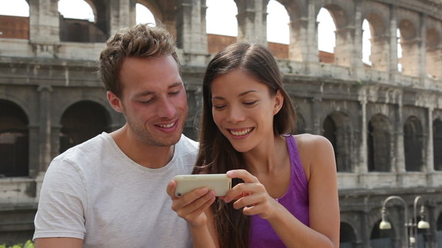 Couple in Rome by Colosseum using smart phone looking at pictures or using travel app in Italy. Happy lovers on honeymoon sightseeing Coliseum. Love and travel concept with multiracial couple.