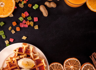 top view of food: waffles, orange, cinnamon, candied fruit on a black and gray background