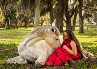 White Azteca horse mare stallion equine lying down in field pasture meadow with young woman girl lady in a red dress gown sitting down looking romantic serene innocent trusting beautiful connected