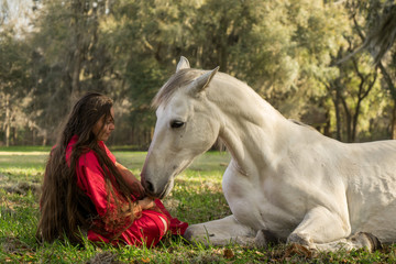 White Azteca horse mare stallion equine lying down in field pasture meadow with young woman girl lady in a red dress gown sitting down looking romantic serene innocent trusting beautiful connected