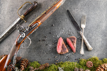 deer or venison steak with antique long gun, cutlery and ingredients like sea salt and pepper, food background for restaurant or hunting loving