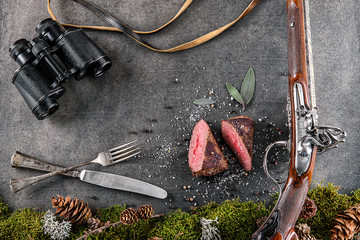 deer or venison steak with antique long gun, cutlery, binocular and ingredients like sea salt, herbs and pepper, food background for restaurant or hunting loving - obrazy, fototapety, plakaty