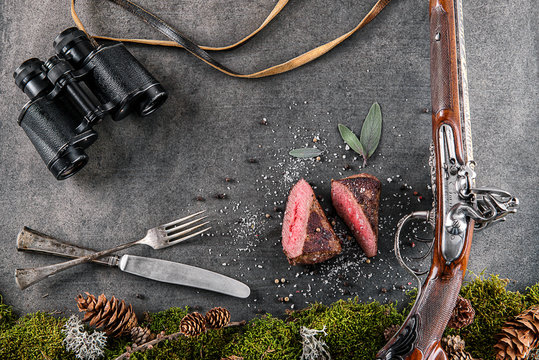 deer or venison steak with antique long gun, cutlery, binocular and ingredients like sea salt, herbs and pepper, food background for restaurant or hunting loving