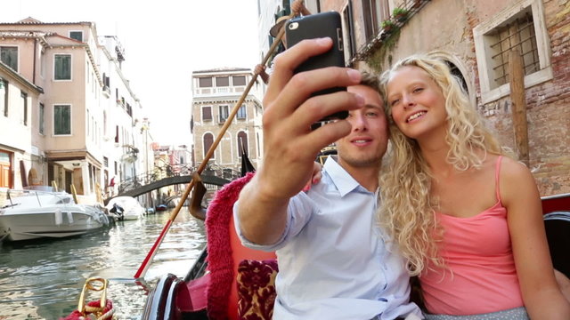 Couple in Venice on Gondole ride romance in boat happy together on travel vacation holidays. Romantic young beautiful couple taking self-portrait sailing in venetian canal in gondola. Italy.