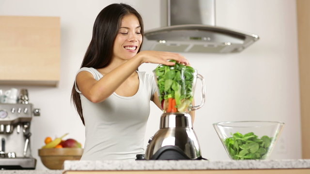 Woman making green vegetable smoothie putting spinach in blender and blending smoothies. Healthy eating lifestyle with young woman preparing drink with spinach, carrots, celery at home in kitchen.