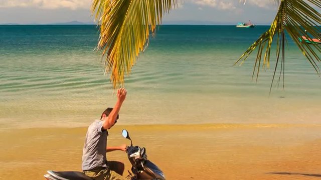 Guy Sits on Motorbike Smiles Touches Palm Branch under Palm