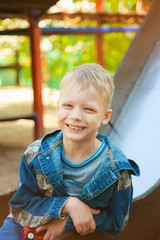 Close up portrait of funny smiling child face. 7 years old kid playing at children playground. Caucasian blond boy dressed in casual denim clothes. Happy kid sitting on slide on spring or summer day.