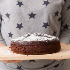 Homemade fresh chocolate birthday cake with white sugar powder on the top in the hands of a woman. A nice cake for a party. Grey background with navy stars and wooden board.