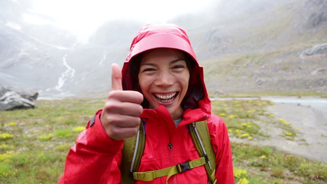 Thumbs up hiking woman happy looking at camera smiling happy and positive on hike in rain in mountain. Multicultural Asian Caucasian female trekking showing success hand sign in Swiss Alps Switzerland