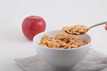 Corn and rice flakes in a spoon and a red apple on a white backg
